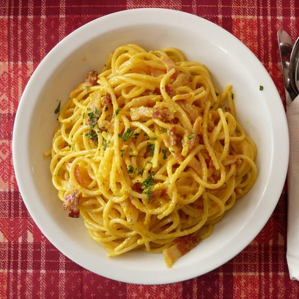 A white bowl of spaghetti carbonara with bacon and parsley, on a red and white checkered tablecloth.